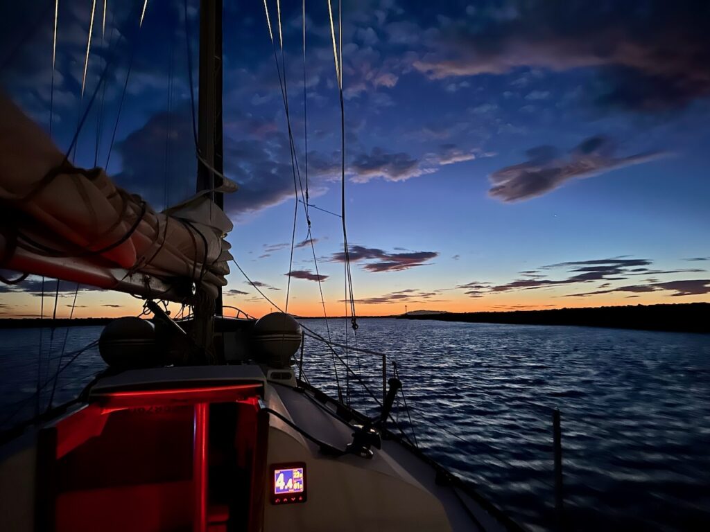 sailboat underway at dusk, red night lights glowing in cabin, scattered clouds in evening lihgt